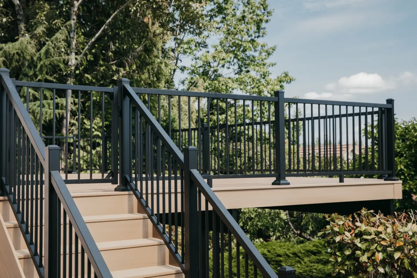 A modern outdoor staircase with black metal deck railing surrounding a raised wooden deck amid green trees and shrubs