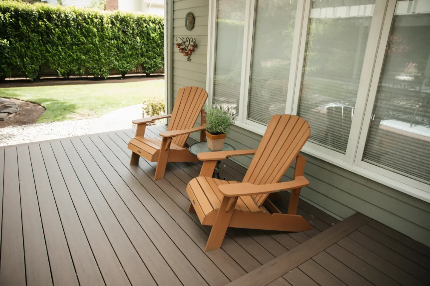 Composite deck with two wooden adirondack chairs potted plants and large windows overlooking manicured backyard with green hedge fence