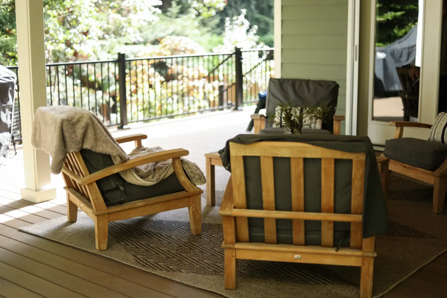Cozy covered porch with wooden lounge chairs cushions and throw blanket on outdoor rug overlooking green trees and black railing