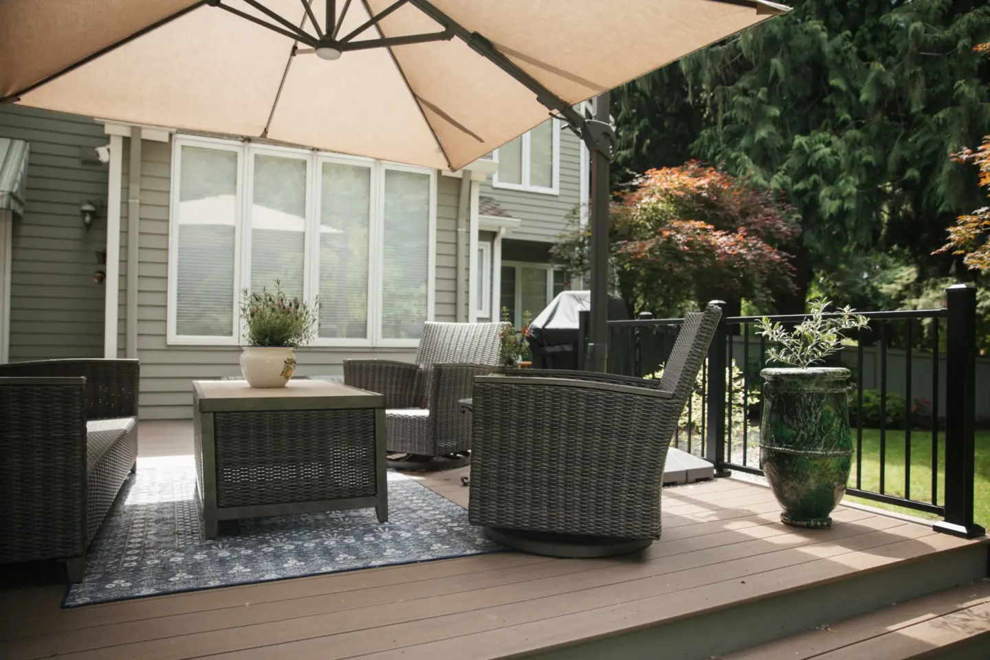 Outdoor patio deck with wicker chairs square coffee table and large beige umbrella next to house and garden railing