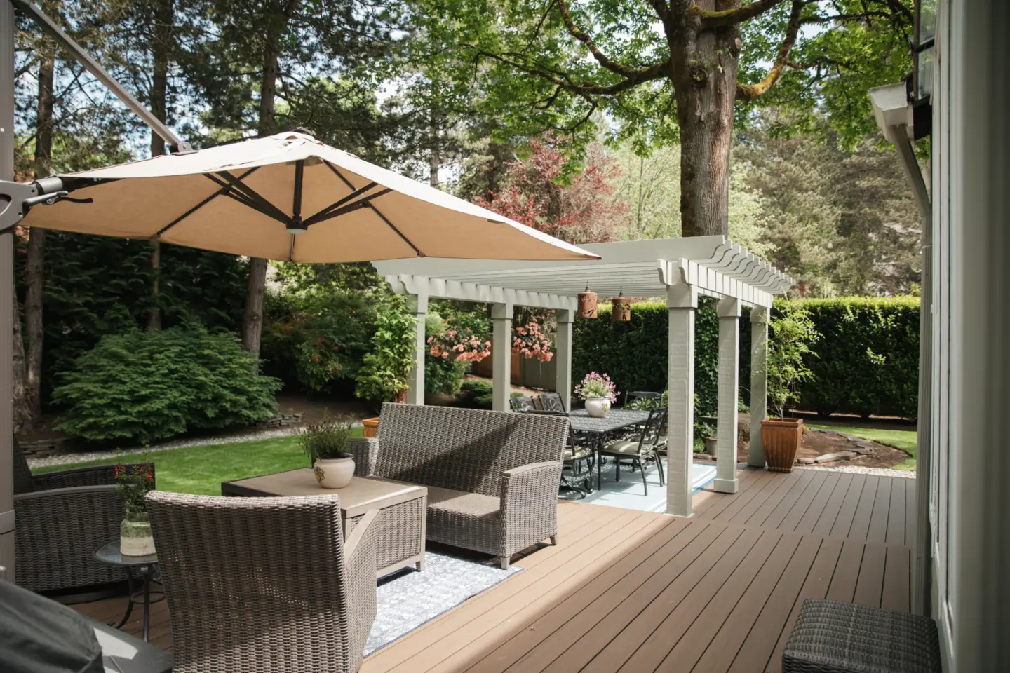 Outdoor wooden deck with wicker patio furniture large beige umbrella and white pergola surrounded by green trees and landscaped garden