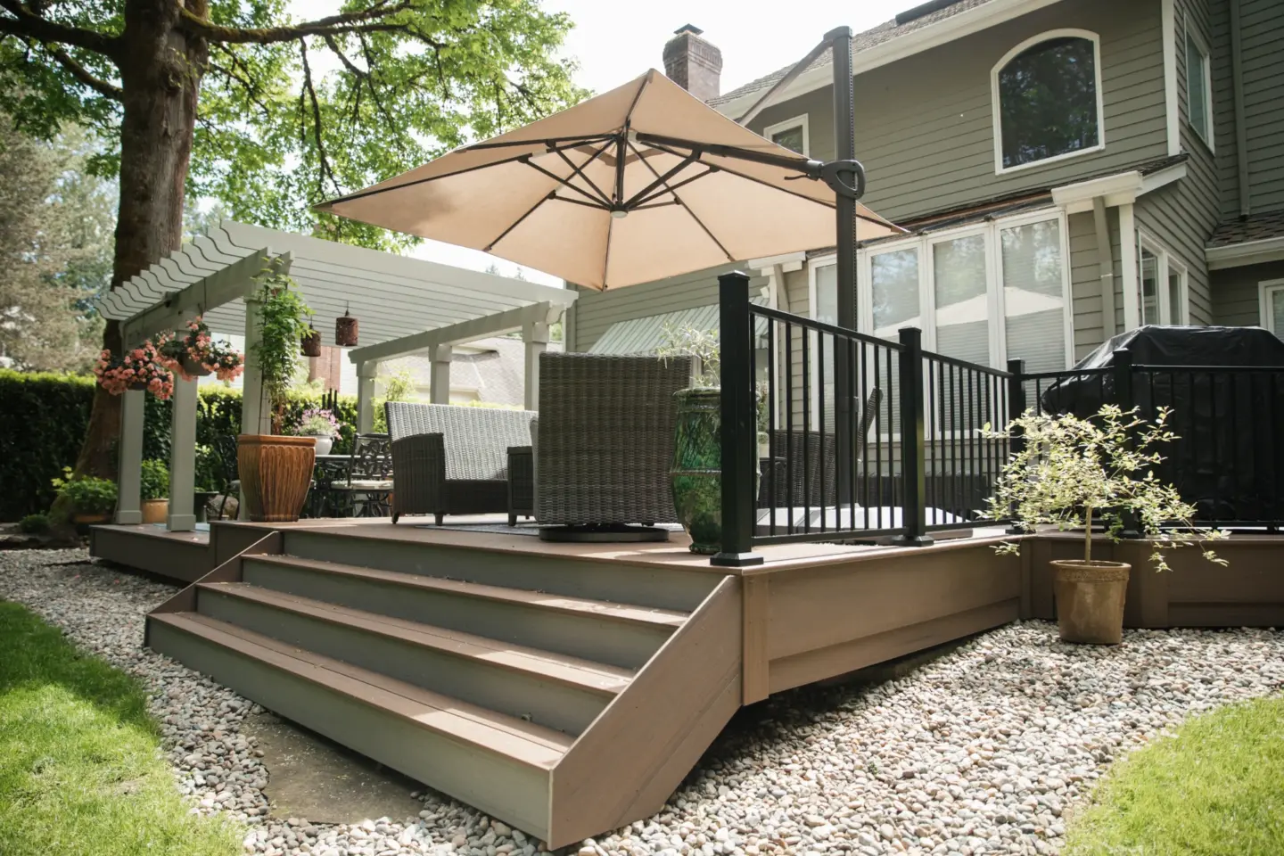 Raised backyard deck with steps wicker patio furniture large beige umbrella black railing and white pergola beside house and garden