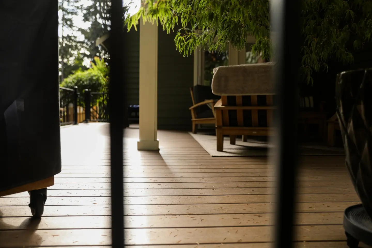 Sunlit wooden porch floor with outdoor seating partially framed by black railings and overhanging green branches